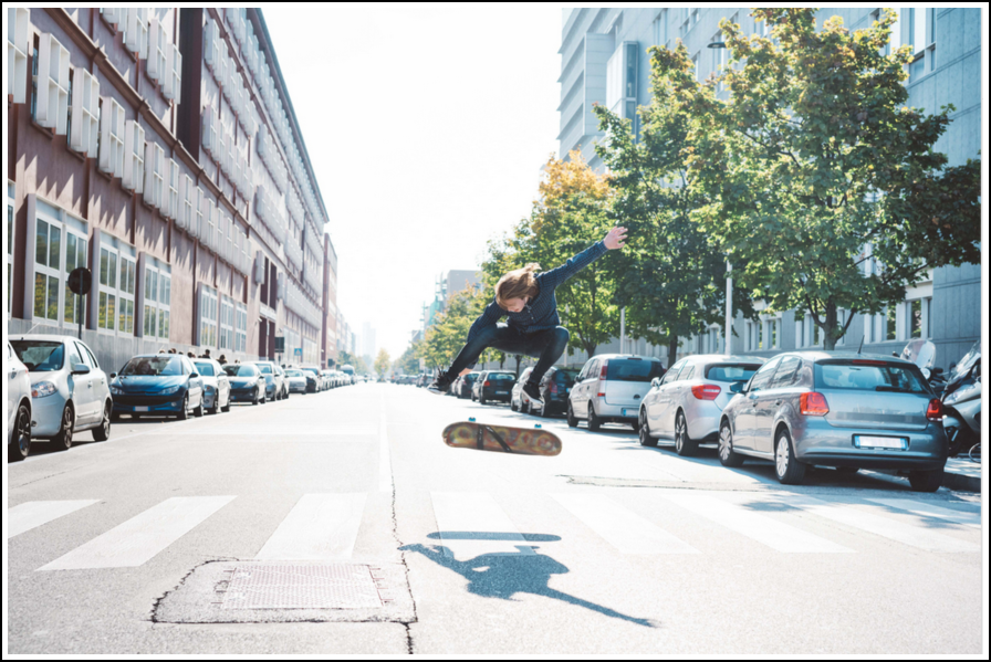 Person, die mitten auf einer Stadtstraße zwischen geparkten Autos einen Stunt auf einem Skateboard macht.