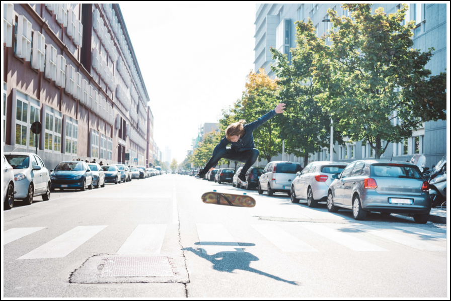 Persona haciendo una acrobacia en una patineta en medio de una calle de la ciudad entre coches aparcados.