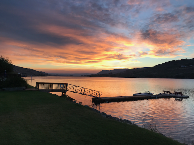 Coucher de soleil sur un lac avec un quai et de petits bateaux, entouré de montagnes.